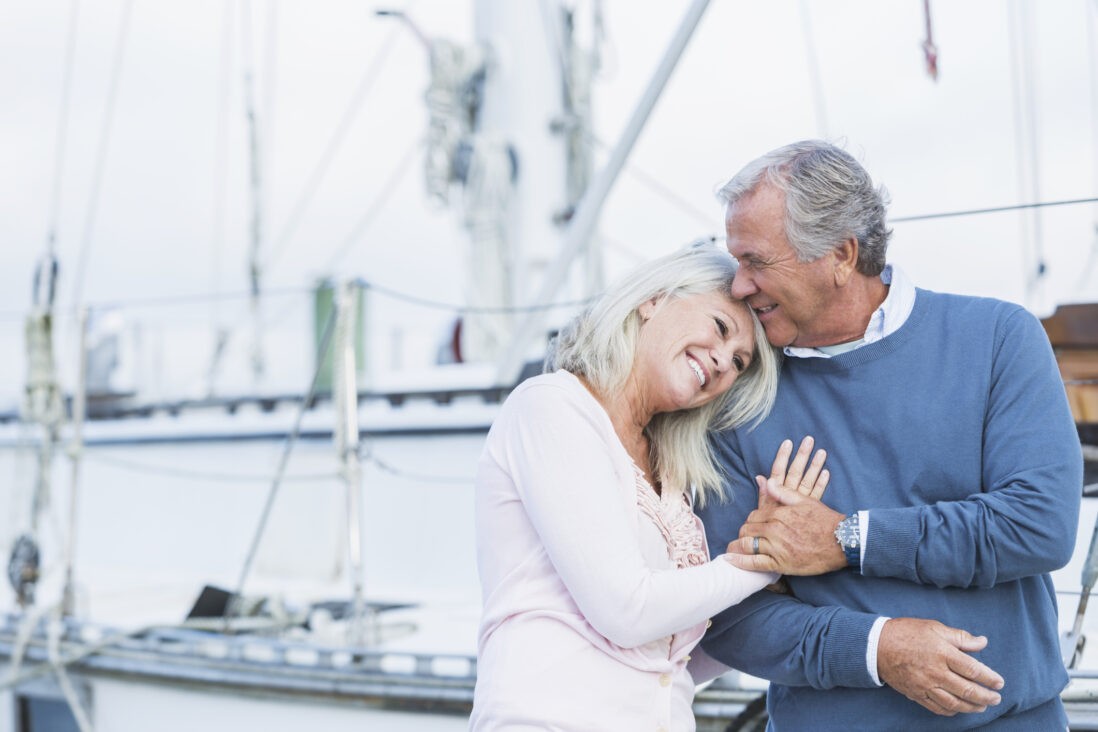 ELDERLY COUPLE HUGGING AND SMILING IN FRONT OF SAILBOATS